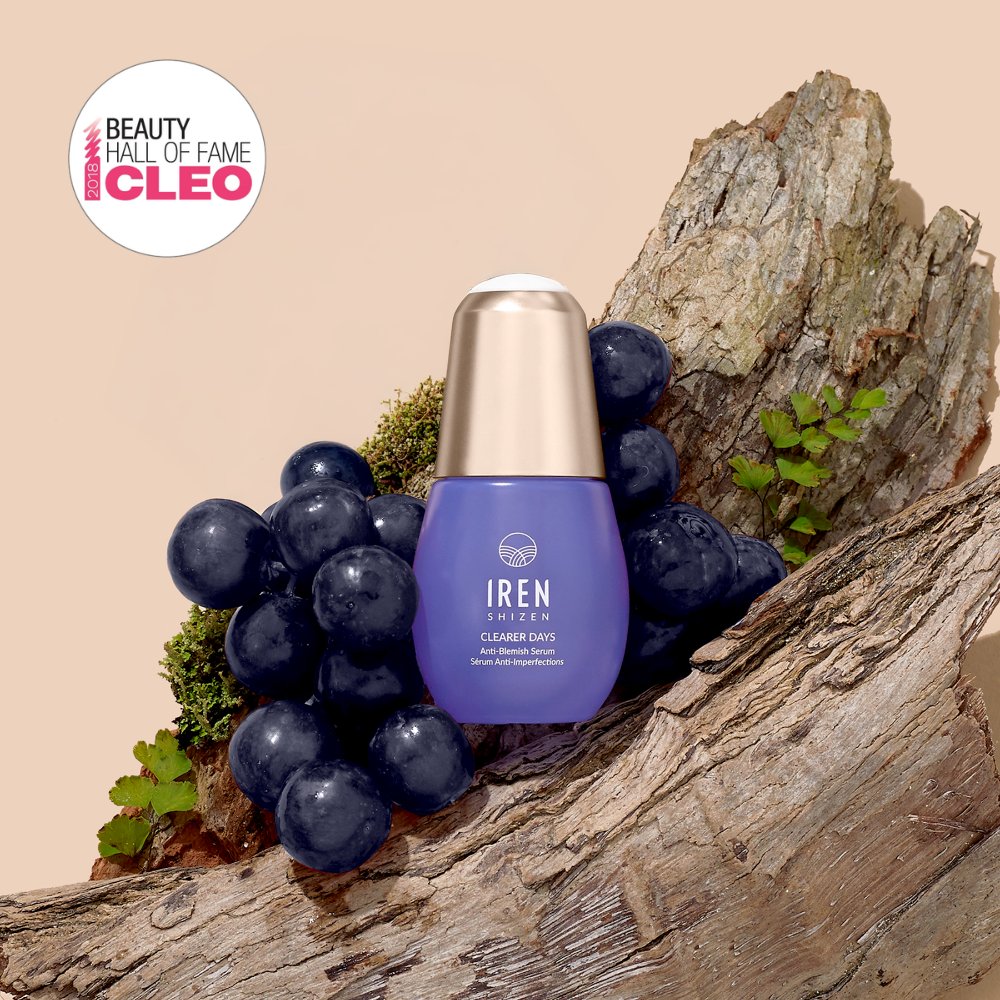 A purple bottle of IREN Shizen CLEARER DAYS Anti-Blemish Serum is displayed among dark grapes and tree bark, featuring a "Cleo Beauty Hall of Fame" badge in the top left corner.