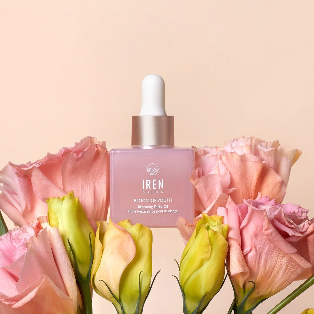 A pink dropper bottle labeled "BLOOM OF YOUTH Restoring Facial Oil" by IREN Shizen sits among pink and yellow flowers against a light beige background, highlighting its lightweight formula that supports skin barrier repair.