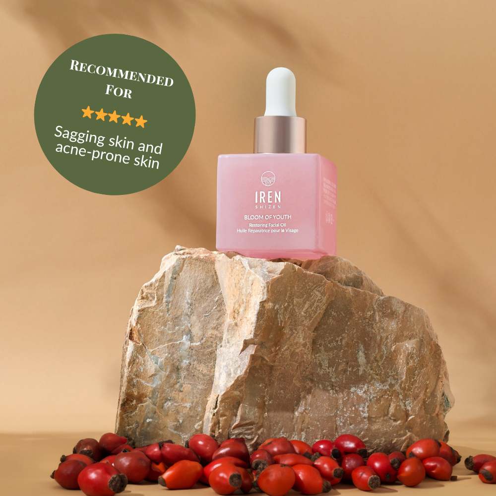 A pink bottle of BLOOM OF YOUTH Restoring Facial Oil by IREN Shizen with a dropper sits on a rock among rosehips; its green label highlights prebiotics and postbiotics for supporting the skin barrier, targeting sagging and acne-prone skin.