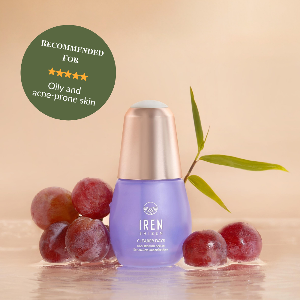 A bottle of IREN Shizen CLEARER DAYS Anti-Blemish Serum sits on a wet surface with red grapes, a green stem, and a badge recommending it for sebum balance and oily, acne-prone skin.