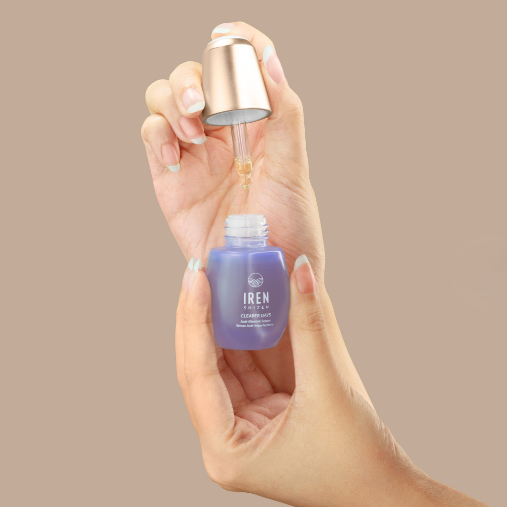 A hand holds a purple bottle of IREN Shizen CLEARER DAYS Anti-Blemish Serum with a dropper, dispensing liquid for sebum balance, against a plain beige background.