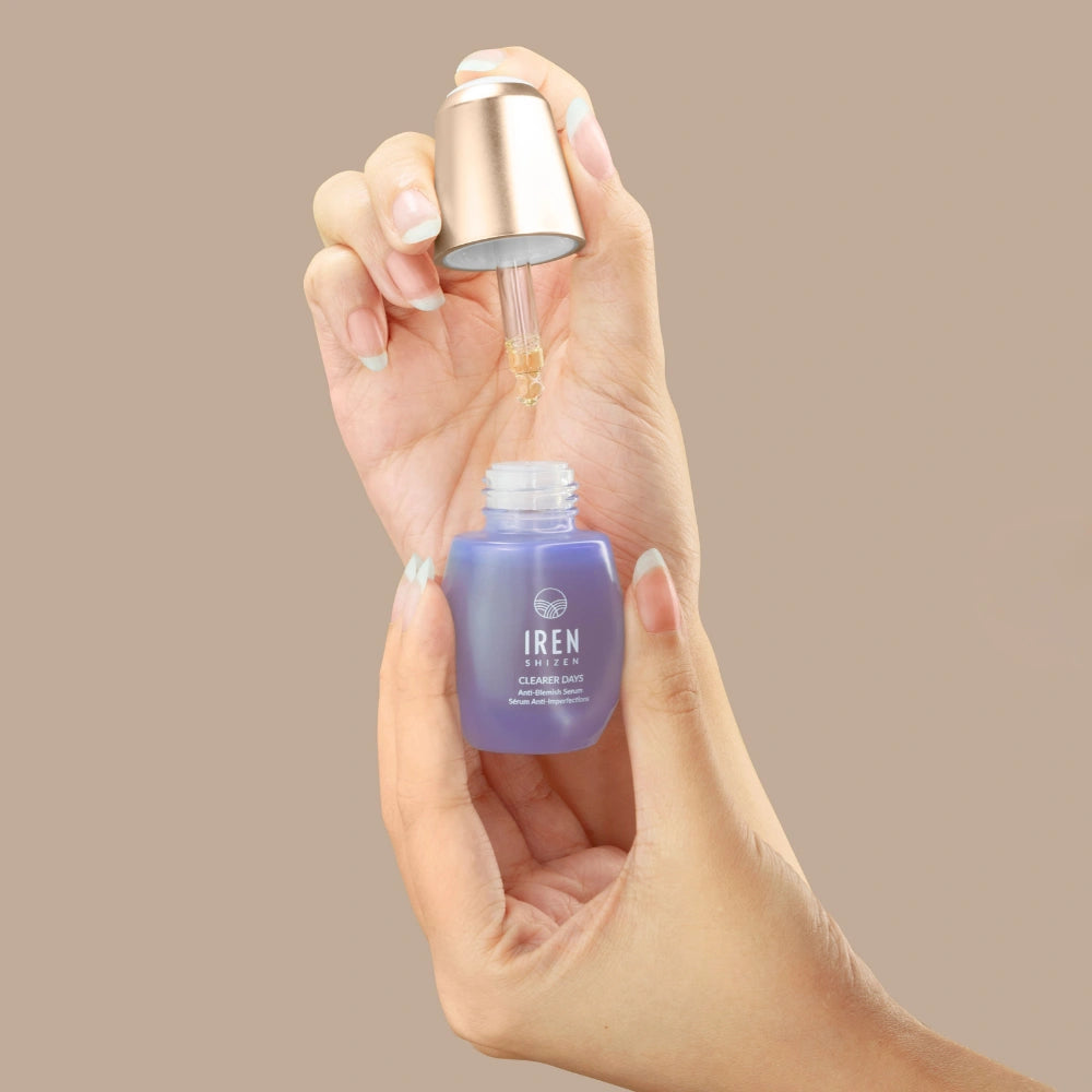 A hand holds a purple IREN Shizen CLEARER DAYS Anti-Blemish Serum bottle while another dispenses the serum with a dropper against a beige background, highlighting its acne treatment and sebum-balancing benefits.