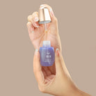 A hand holds a purple IREN Shizen CLEARER DAYS Anti-Blemish Serum bottle while another dispenses the serum with a dropper against a beige background, highlighting its acne treatment and sebum-balancing benefits.