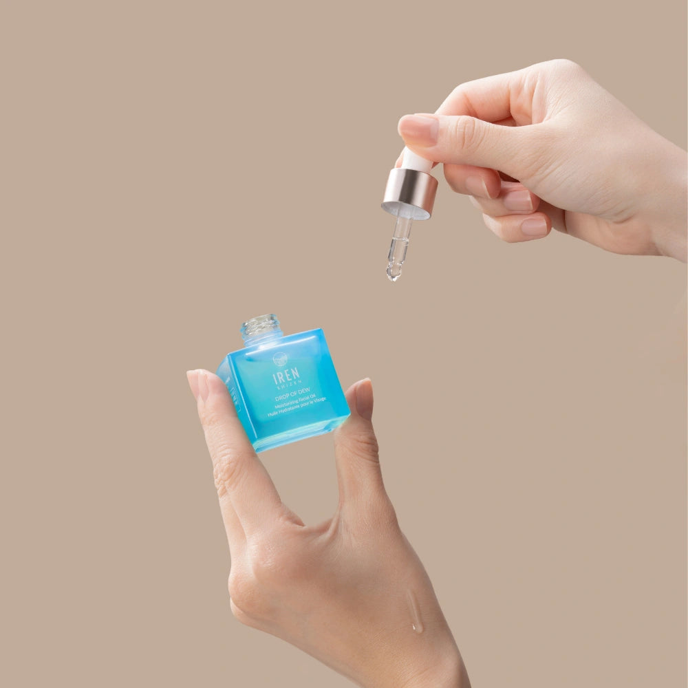 A hand holds IREN Shizen DROP OF DEW Moisturizing Facial Oil, dispensing the multi-tasking, cold-pressed oil for skin barrier repair with a dropper, set against a beige background. #all-groups