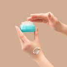 A hand holds an open jar of IREN Shizen FREEZE THE YOUTH Hydra Firming Cream while the other dips in; a smear of this hydrating, age-defying moisturizer is visible on the wrist against a beige background.