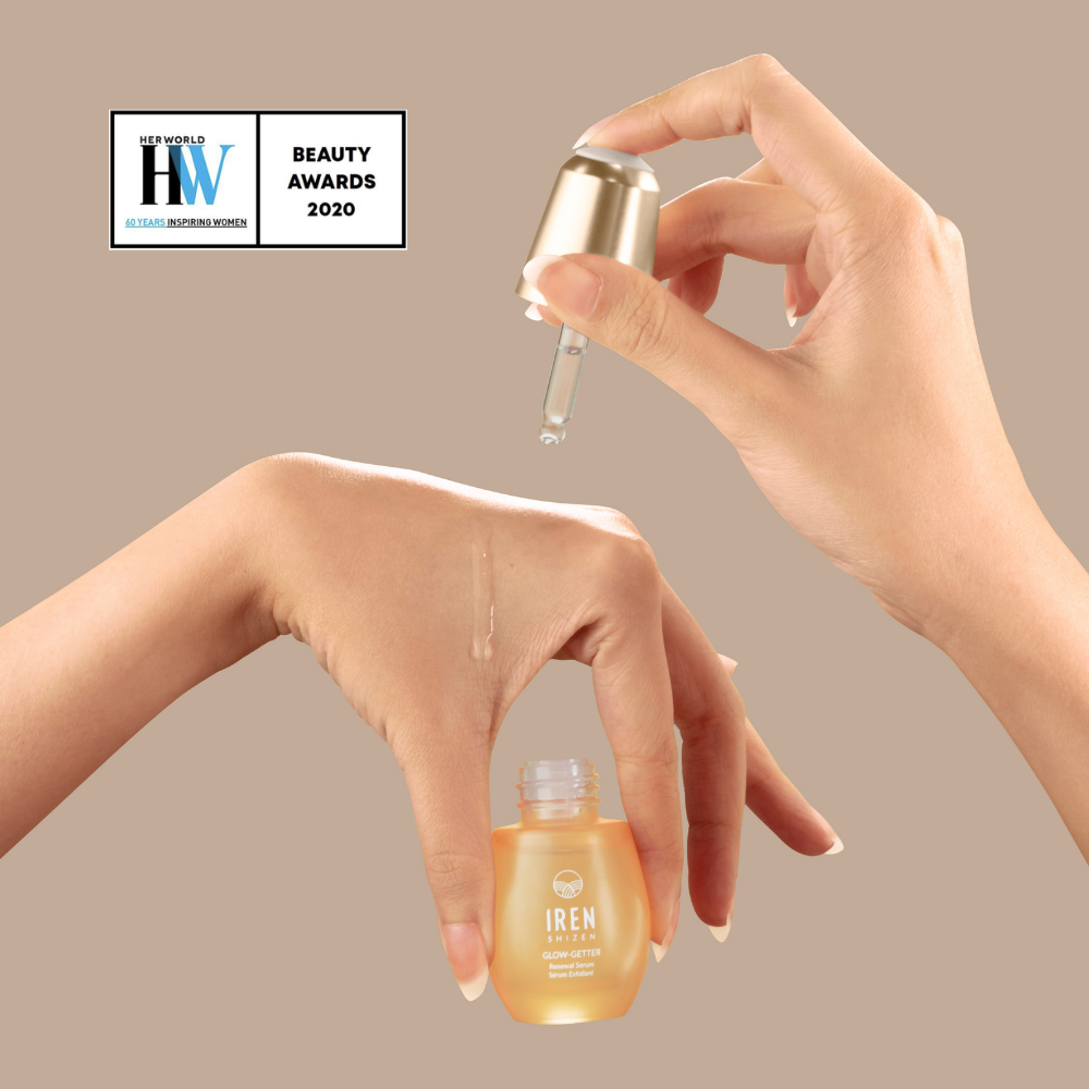 A hand applies GLOW-GETTER Renewal Serum from IREN Shizen onto the back of another hand using a dropper; a small bottle labeled “IREN” sits below with a beauty awards badge. Ideal for those seeking to exfoliate and renew their skin's natural glow.
