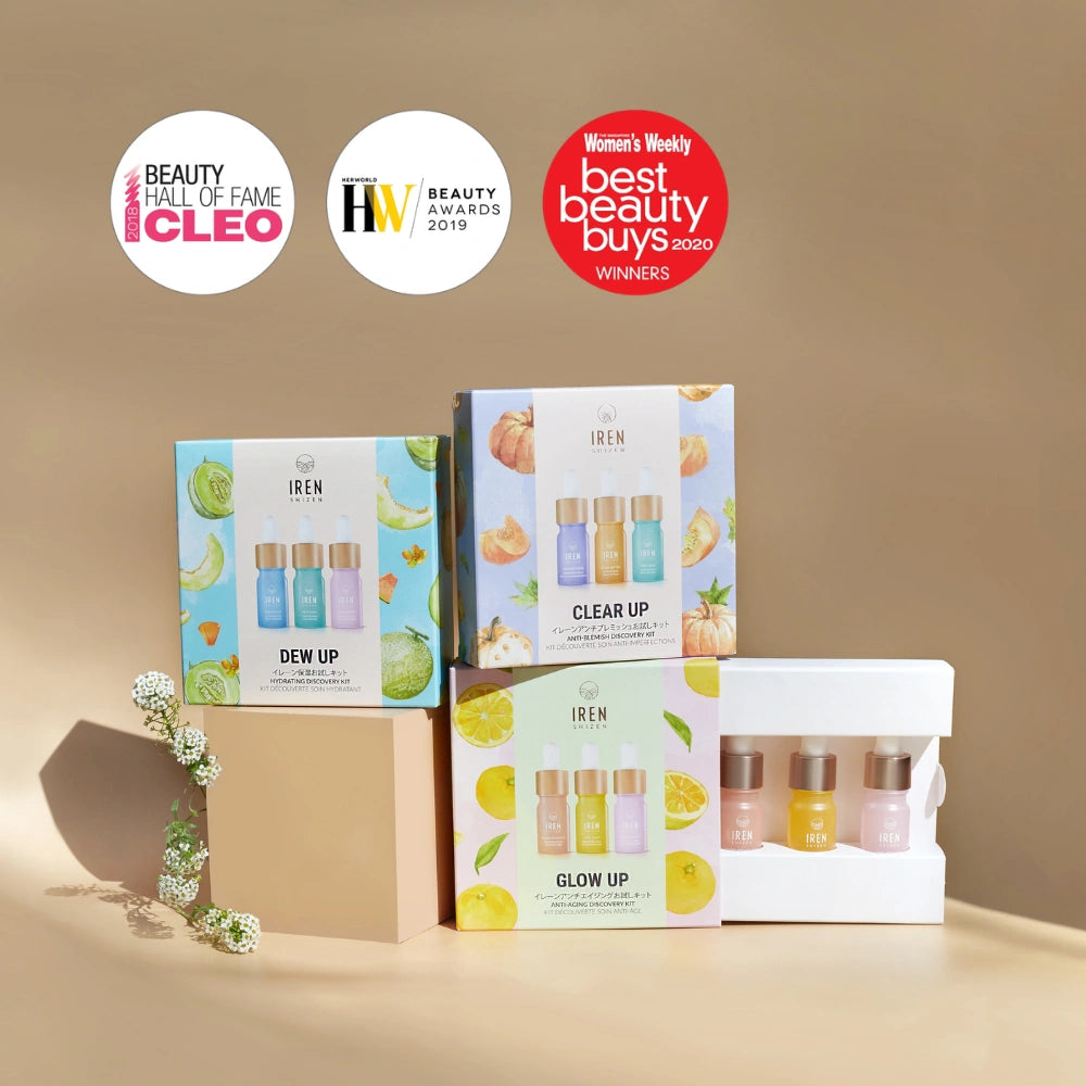 The IREN Shizen GLOW UP Anti-Aging Discovery Kit and a trio of brightening serums are displayed on a beige surface with award badges in the background.