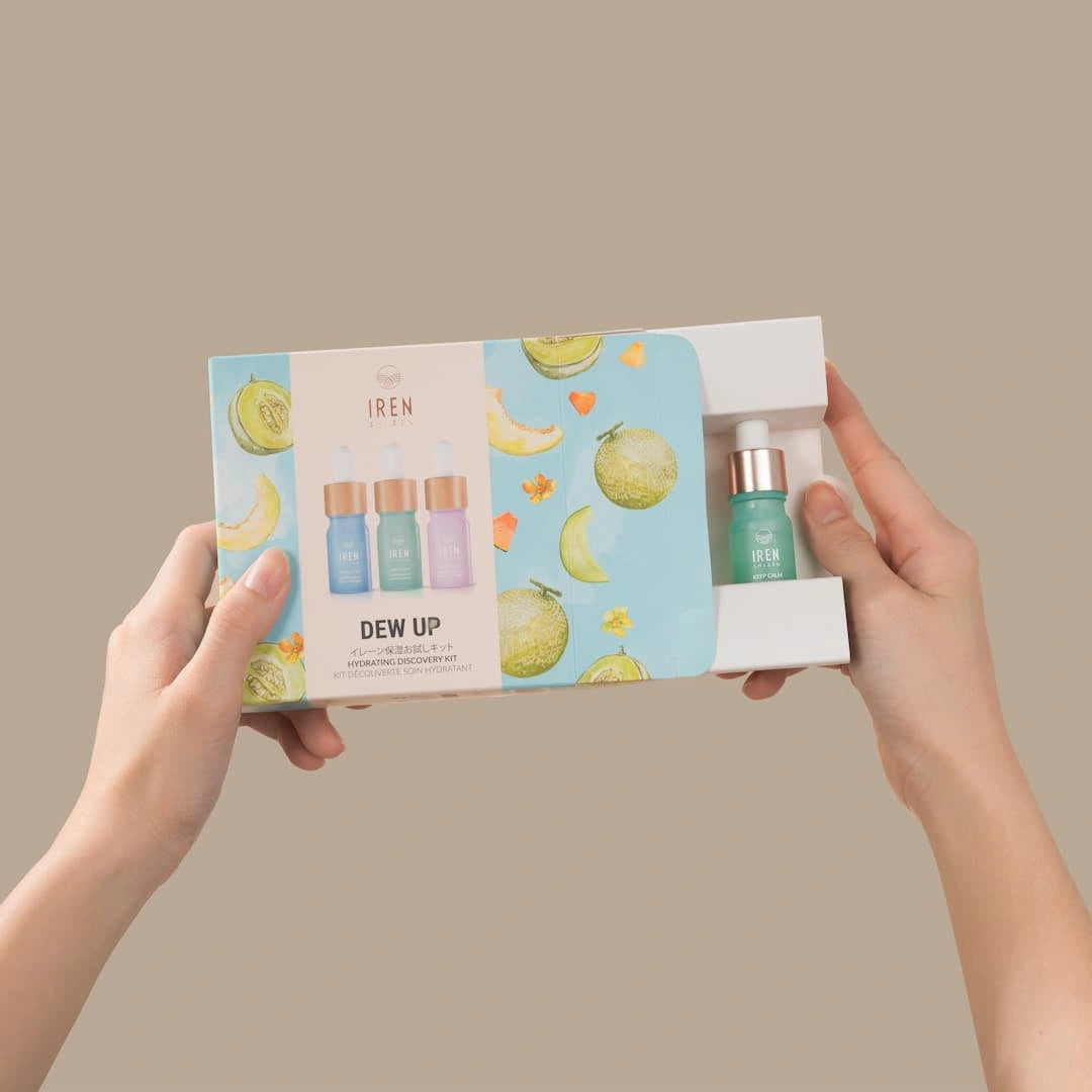 Hands hold the DEW UP Hydrating Discovery Kit by IREN Shizen, with colorful melon and flower illustrations. The partially opened box reveals a small dropper bottle of hydrating serum, perfect for achieving dewy skin.