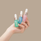 A hand holds three dropper bottles from the IREN Shizen DEW UP Hydrating Discovery Kit in green, blue, and pink, set against a plain beige background.