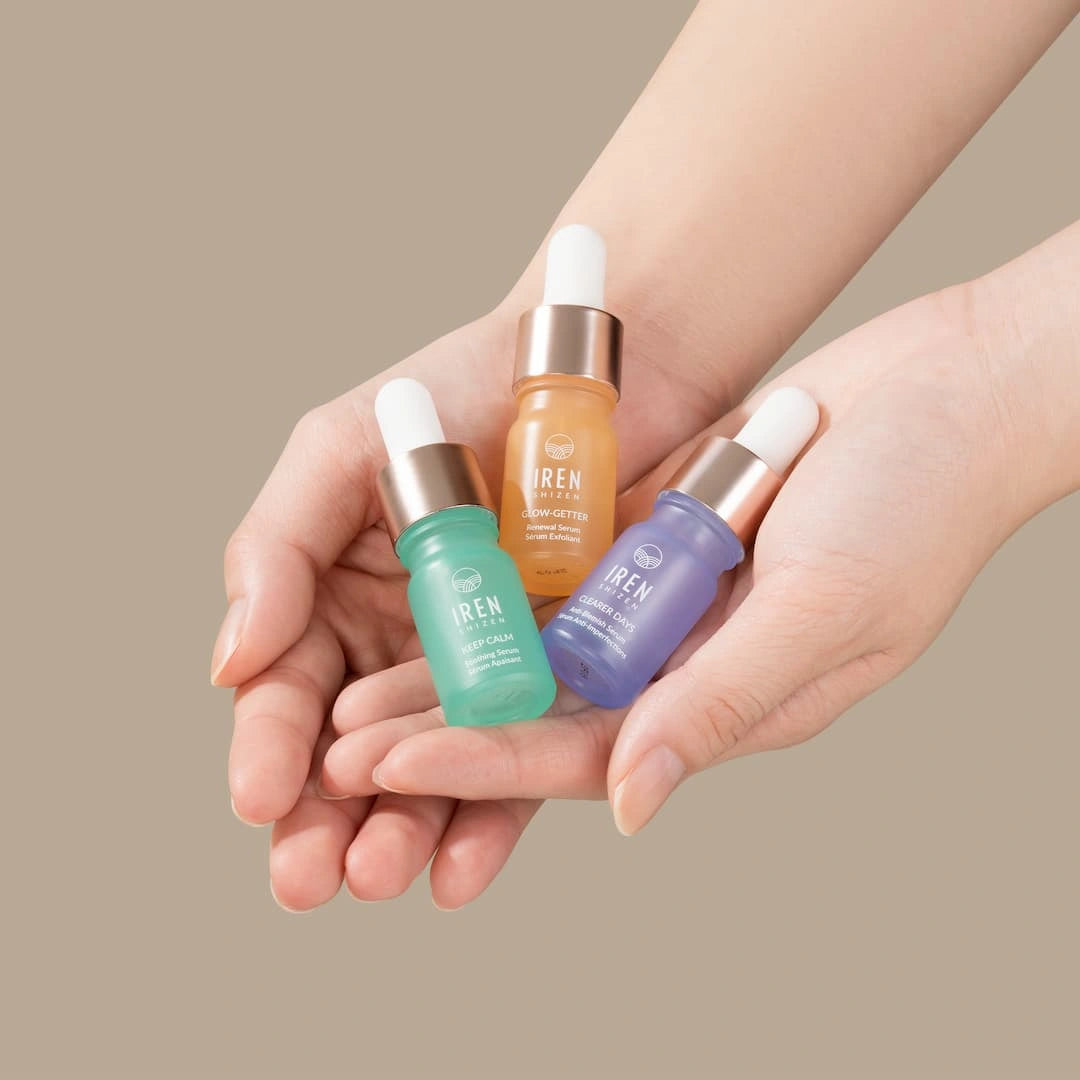 A pair of hands holds three dropper bottles from the CLEAR UP Anti-Blemish Discovery Kit by IREN Shizen, featuring serums in green, orange, and purple—each infused with Japanese superfruits—against a neutral background.