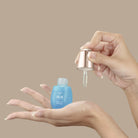 A hand holds an open blue QUENCH-UP Hydrating Serum bottle by IREN Shizen while another dispenses hyaluronic acid with a dropper onto the palm, promising plump skin against a beige background.