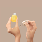 A person holds a small yellow bottle of IREN Shizen STAR LIGHT Brightening Serum in one hand and a dropper in the other, ready to target hyperpigmentation, against a plain beige background.