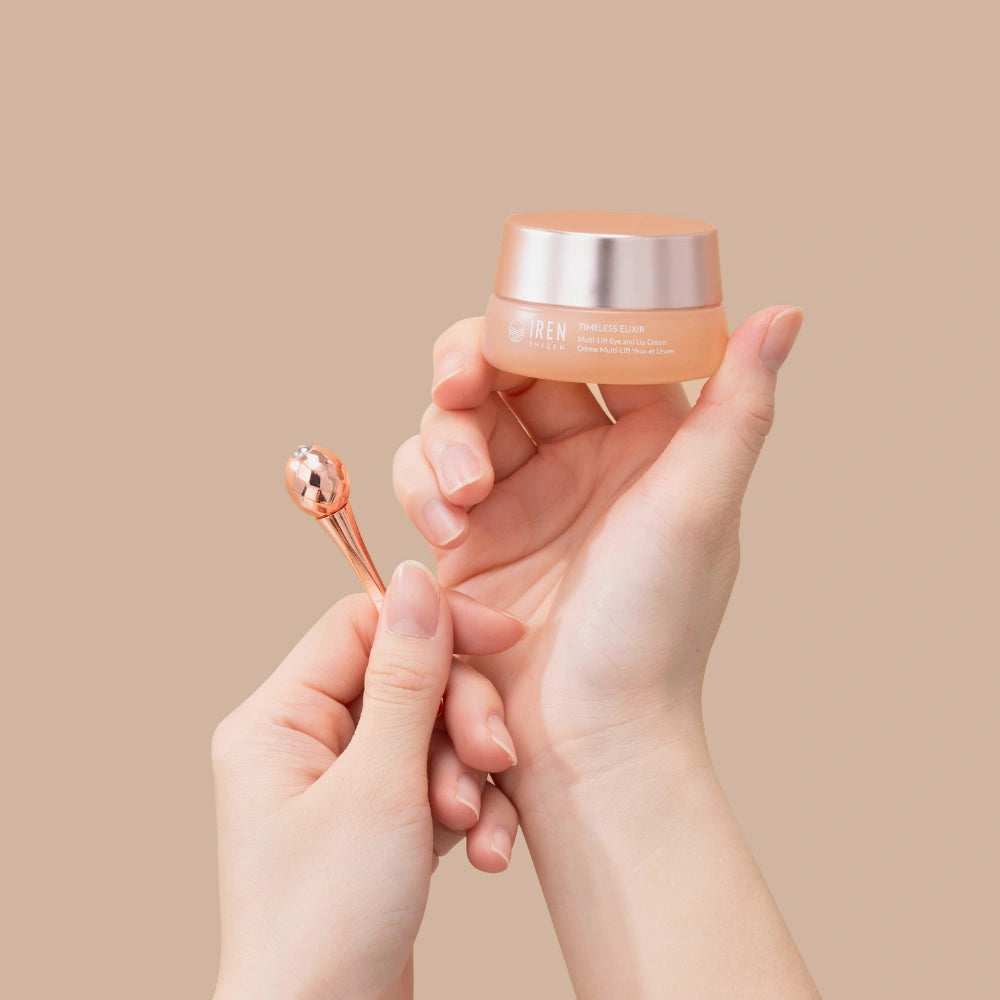 A person holds a jar of IREN Shizen TIMELESS ELIXIR Multi-Lift Eye and Lip Cream in one hand and a rose gold applicator in the other, set against a beige background—an elegant solution for treating dark circles.