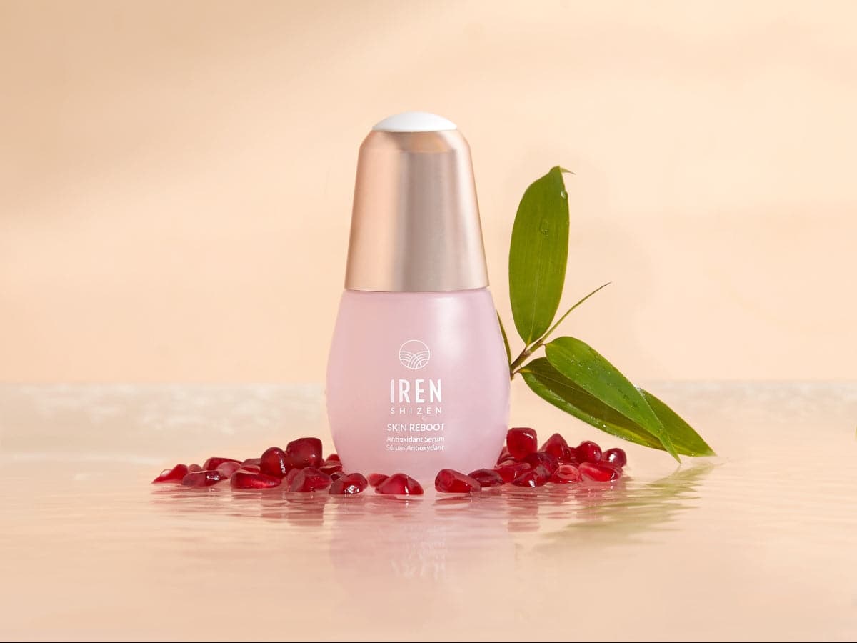 A pink bottle labeled "IREN Shizen APAC TIMELESS ELIXIR Anti-Aging Eye Skincare Kit" sits elegantly on a glossy surface, surrounded by pomegranate seeds and a green leaf, epitomizing the essence of premium anti-aging serums.