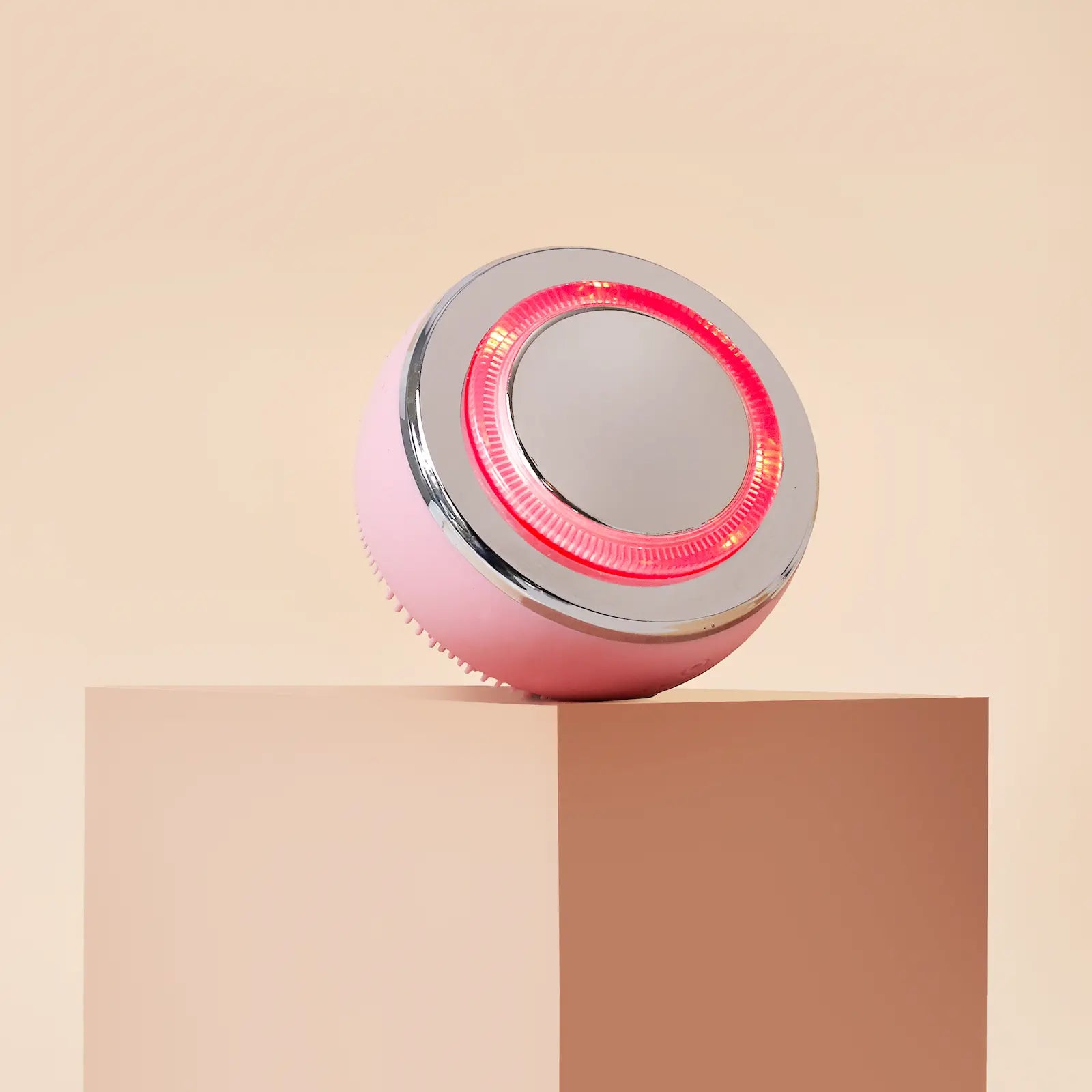 The IREN Shizen GLOW ON THE GO Travel Light Kit, a round pink and silver facial cleansing brush with red LED light therapy, rests on the edge of a beige rectangular block against a neutral background.