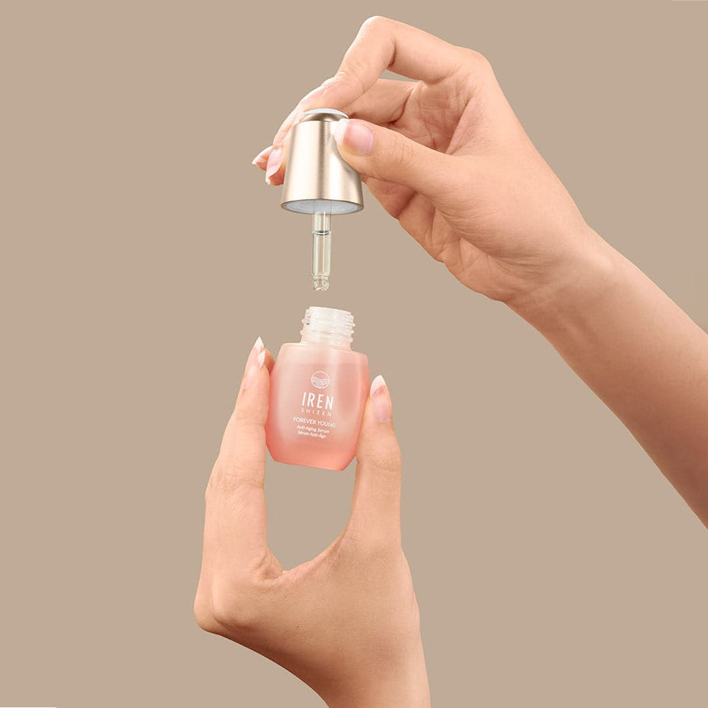 A hand is holding a bottle of IREN Shizen customised FOREVER YOUNG Anti-Aging Serum on a beige background.